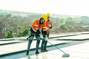 Facility maintenance workers in hi-vis orange jackets and yellow hard hats cleaning rooftop solar panels with safety harnesses, overlooking a housing estate.