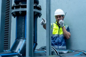 Worker in hi vis jacket and hard hat using a two way radio beside water treatment pipework.