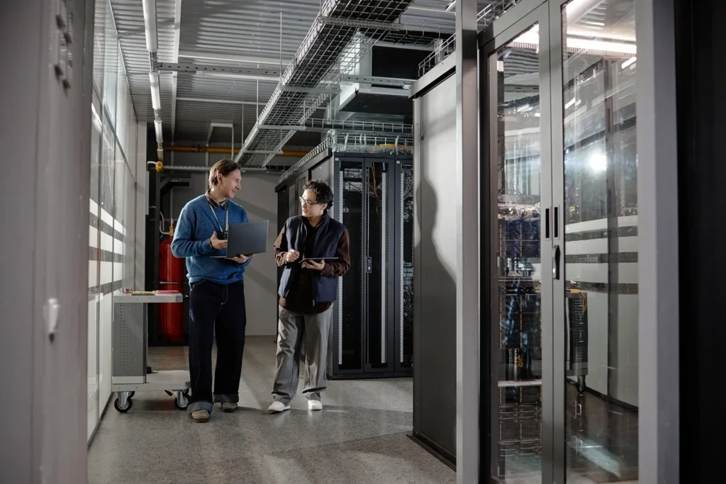 Data centre engineers using a two way radio and laptop while working beside server racks in a technical facility.